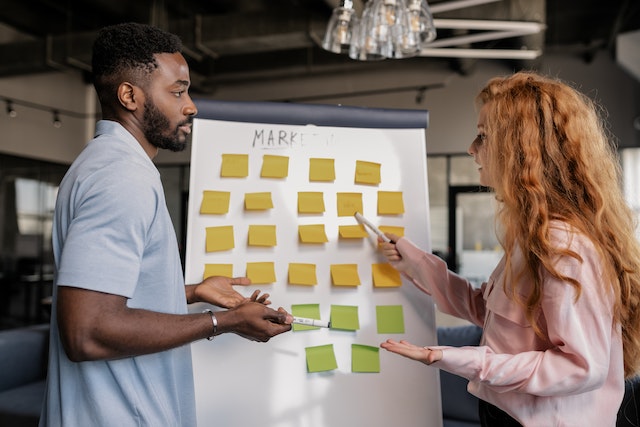 two people discussing a whiteboard full of sticky notes that says "marketing" at the top